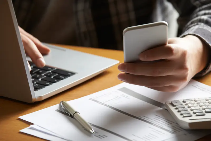Hände an Laptop und Smartphone, daneben Unterlagen, Kugelschreiber und Taschenrechner